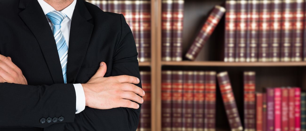 Midsection of lawyer with arms crossed standing against books in shelves